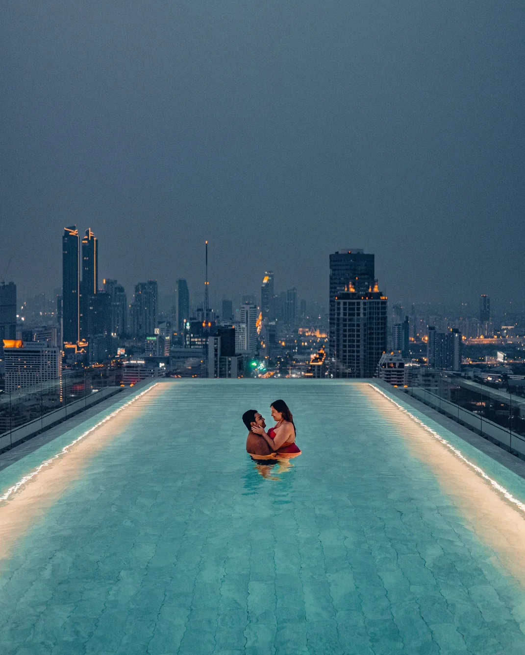 Pao y Andrew en una piscina infinita con la ciudad iluminada al fondo.